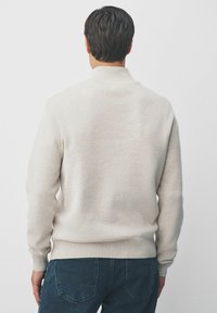 Man met kort, donker haar, gekleed in een lichtbeige geribde trui en donkerblauwe jeans, met zijn rug naar de camera gekeerd tegen een effen witte achtergrond.