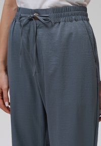 Svetle šedé kalhoty s elastickým pasem a detaily stahovací šňůrky. Hladký materiál, boční kapsy a jemně nařasený pas.