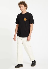 Sort T-shirt med korte ærmer, der har et rundt orange grafisk tryk, parret med cremefarvede bukser og sorte sko. Enkel, afslappet design.