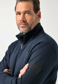 Marineblauwe sweatvest met ritssluiting van zacht materiaal, voorzien van een hoge kraag, een subtiel logo op de borst en een gestructureerde zwarte patch op de mouw.