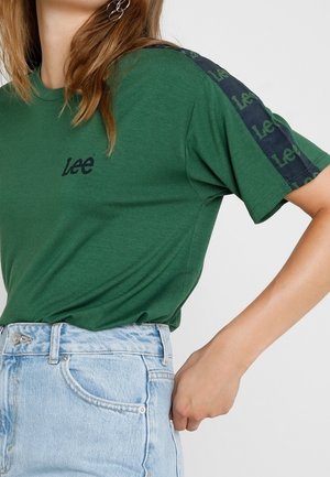Green cotton t-shirt with short sleeves, featuring a black logo and patterned shoulder accents. Paired with light blue high-waisted denim.