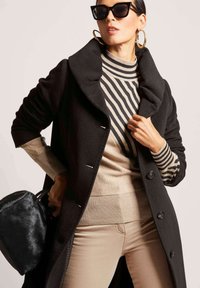 Zwarte wollen jas met een grote kraag, gecombineerd met een beige en zwart gestreepte sweater. Accessoires zijn onder andere een kleine zwarte bont handtas en oversized zonnebril.