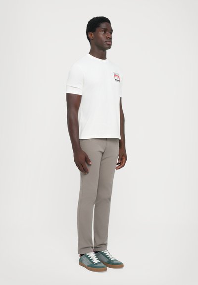 Vit kortärmad t-shirt med grafisk lapp, parat med ljusbeige slim-fit byxor och grön-blå sneakers med vita snören och bruna sulor.