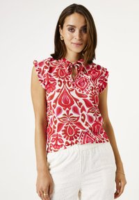 Blusa de manga corta con un lazo en el cuello, que presenta un estampado floral en rojo y rosa sobre un fondo blanco. Las mangas con volantes añaden un toque de detalle.