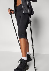 Leggings capri neri con un design texture vicino all'orlo, abbinati a scarpe da trekking nere e bastoni da trekking. La giacca ha una texture grigia a rete.
