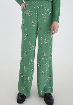 Pantalons larges fleuris verts avec une texture douce, ornés de motifs de fleurs blanches et orange, associés à des chaussures noires à semelle épaisse.