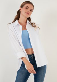 White button-up shirt made of lightweight fabric, featuring long sleeves, relaxed fit, and a crisp collar, layered over a blue cropped top.