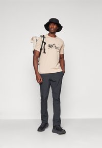 Beżowa koszulka z grafiką gór, w połączeniu z ciemnoszarymi spodniami i czarnymi butami trekkingowymi. Czarne kapelusz typu bucket i biały plecak dopełniają stylizację.