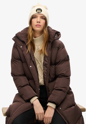 Bruine pufferjas met een hoge kraag en zilveren knopen, gelaagd over een crèmekleurige gebreide trui. Beige beanie met een nep bont pompon en logo.