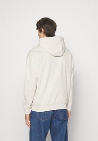 Hoodie beige clair avec une texture à motifs, doté d'une capuche avec cordon de serrage et de poignets côtelés. Associé à un jean en denim bleu.