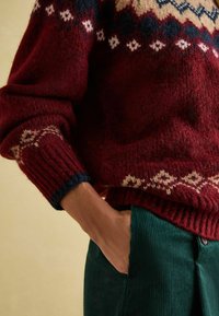 Roter und cremefarbener gemusterter Strickpullover mit gerippten Manschetten und Saum, kombiniert mit dunkelgrünen Cordhosen mit Seitentaschen.