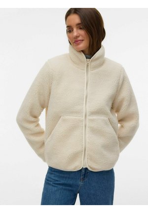 Kvinne iført en kremfarget sherpa fleecejakke med glidelås og høy krage, samt blå jeans, som ser ned med hendene bak ryggen.