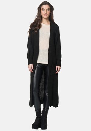 Lungo cardigan nero lavorato a maglia con apertura frontale, polsini a coste e tasche, abbinato a una camicia leggera e leggings neri lucidi.