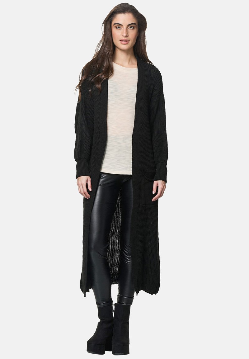 Lång, stickad svart cardigan med öppen framsida, ribbade manschetter och fickor, i kombination med en ljus skjorta och glänsande svarta leggings.