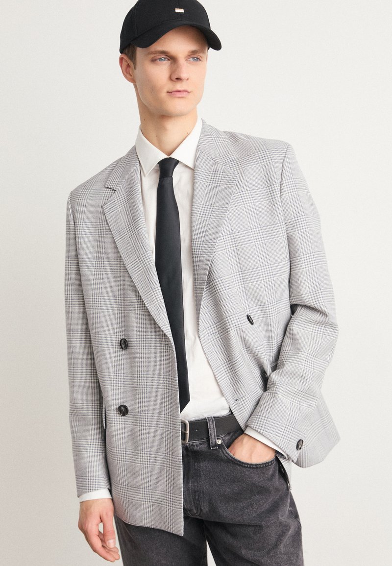 Double-breasted blazer in gray and white check pattern, featuring large black buttons, worn over a white shirt and black tie.
