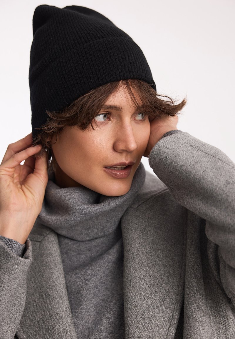 Czarna, prążkowana czapka beanie na głowie modela, zestawiona z szarym swetrem z golfem i płaszczem, prezentująca miękkie tekstury i zbliżenie na twarz.