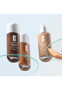 Trois flacons de Clinique Even Better Clinical Vitamin Makeup SPF 50 en différentes teintes, dont un avec une pompe et un échantillon de liquide.