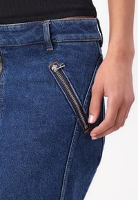 Blå denimjeans har en særpreget sidelomme med glidelås og metall detaljer. Teksturen er glatt, og stoffet viser en klassisk denimveving.