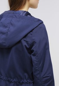 Veste à capuche bleu marine avec une texture lisse, taille élastique et détails simples de couture. Le dos présente une coupe décontractée.