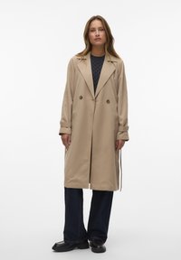 Cappotto trench beige doppiopetto con ampi risvolti, tasche laterali e dettaglio cinturino. Indossato sopra un top testurizzato blu navy e jeans scuri.