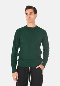 Maglione a coste verde scuro con collo rotondo, maniche lunghe e tessuto testurizzato, abbinato a pantaloni neri con dettagli regolabili.