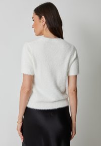 Kobieta z długimi brązowymi włosami ubrana w biały puszysty sweter z krótkim rękawem i czarną satynową spódnicę, odwrócona tyłem na tle jednolitego szarego tła.