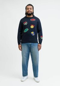 Navy sweatshirt featuring colorful embroidered holiday-themed designs, including gifts and festive text, paired with light blue jeans and white shoes.
