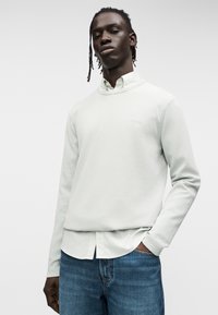 Ανοιχτό γκρι πουλόβερ με ριμπ ύφανση, στρογγυλή λαιμόκοψη και διακριτικό λογότυπο. Φορεμένο πάνω από ένα λευκό πουκάμισο με γιακά, σε συνδυασμό με μπλε τζιν.