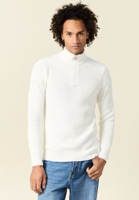 Maglione bianco a coste con colletto alto e chiusura con bottoni. Indossato con jeans in denim blu, evidenziando una forma aderente e un tessuto texturizzato.