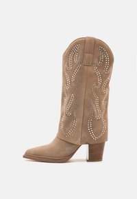 Botas de vaquero de ante marrón claro con punta afilada y tacón cuadrado, que presentan patrones de llamas decorativas con tachuelas en el eje.
