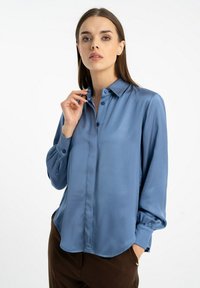 Satinowa bluzka w stonowanym niebieskim kolorze z kołnierzykiem zapinanym na guziki, długimi rękawami i zaokrąglonym dołem. Posiada gładką fakturę i subtelny połysk.