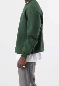 Persona che indossa una felpa verde scuro, una maglietta bianca, pantaloni grigi con strisce laterali e un anello d'argento sulla mano sinistra, su uno sfondo bianco.