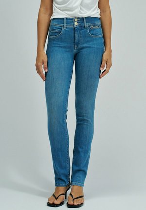 Femme portant un jean slim bleu avec fermeture double bouton et des sandales noires à talons hauts à lanières, debout devant un fond uni.