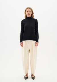 Maglione nero a collo alto con orlo a costine, indossato con pantaloni plissettati crema e ballerine nere a punta, su uno sfondo neutro.