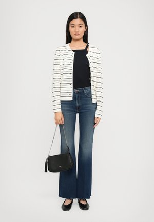 Femme portant un cardigan rayé blanc, un haut noir, un jean évasé bleu, des ballerines noires et tenant un sac à bandoulière noir avec une chaîne argentée.