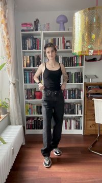 Fekete trikó és sötét farmernadrág. Az illető színes sportcipőt visel. A háttérben egy könyvespolc és egy dekoratív lámpa látható.