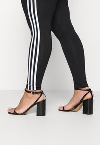 Svarta klacksandaler med blanka detaljer, tunna remmar runt vristen och klack i blockmodell. Bärs med svarta leggings med vita ränder.