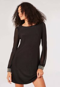 Schwarzes Kleid mit langen, transparenten Ärmeln, rundem Halsausschnitt und verzierten Manschetten mit metallischen Akzenten. Weicher, glatter Stoff mit lockerer Passform.