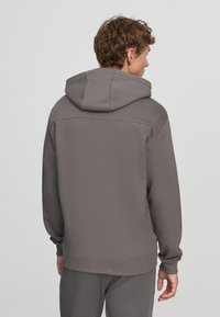 Felpa grigia con cappuccio, maniche lunghe e polsini a coste. Presenta una texture liscia e un design minimalista con un sottile dettaglio sulla cucitura posteriore.