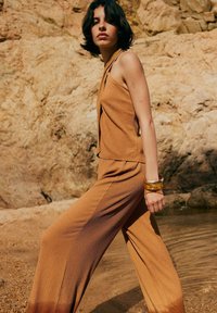 Bruine geribbelde jumpsuit met wijde pijpen en een halterhals, gecombineerd met twee doorzichtige armbanden. Textuurstof tegen een rotsachtige achtergrond.