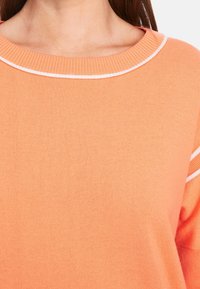 Pull à encolure ronde de couleur orange clair, avec des surpiqûres blanches d'accent le long du col. Texture douce et tricot côtelé.