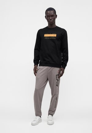 Homme portant un t-shirt à manches longues noir avec le logo orange-jaune EA7 Emporio Armani et un pantalon gris avec le logo EA7 noir le long de la jambe.