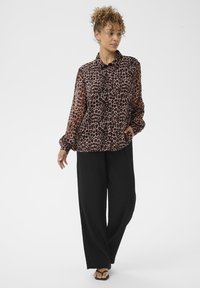 Moteris su garbanotais plaukais, vilkinti ilgomis rankovėmis leopardų rašto bliusone, laisvomis juodomis kelnėmis ir juodomis atviromis basutėmis, stovi prieš baltą foną.