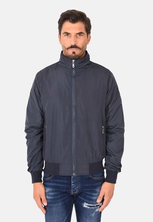 Uomo con capelli scuri e baffi, indossa una giacca bomber blu navy con cerniera e jeans blu strappati, in piedi davanti a uno sfondo chiaro e semplice.