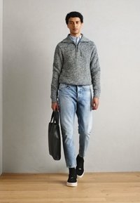 Pull en maille gris, jeans déchirés bleu clair, baskets noires avec semelles blanches, et un sac fourre-tout noir. Debout devant un fond neutre.
