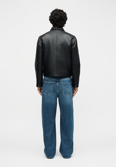 Άνδρας με σγουρά σκούρα μαλλιά, φορώντας μαύρο δερμάτινο μπουφάν και φαρδιά μπλε τζιν, στέκεται με την πλάτη προς το μέρος του θεατή μπροστά σε ένα απλό φόντο.