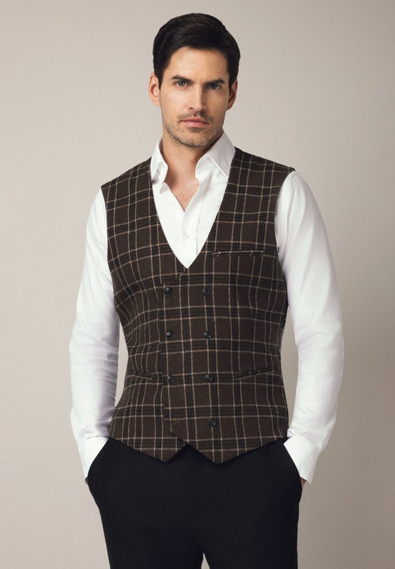Brązowa kamizelka w kratę z podwójnym zapięciem, klapami z wycięciem i jedną kieszenią na piersi, noszona na białej koszuli z czarnymi spodniami.