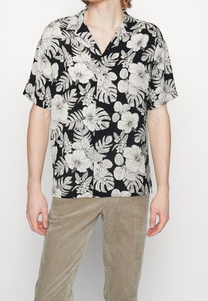 Homme portant une chemise noire à manches courtes avec un imprimé floral tropical blanc et un pantalon en velours côtelé beige, debout devant un fond uni.