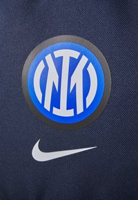 „Nike“ logotipas ant tamsiai mėlynos, tekstūruotos medžiagos su apvaliu „Inter Milan“ emblematu mėlynos ir baltos spalvos centre.