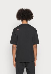 Černé bavlněné tričko s uvolněným střihem, krátkými rukávy a růžovým logem Jumpman na horní části zad. Hladká textura.